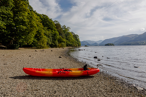 Empty orange and red kayak, on the beach near the lake, mountains in the background and foliage.