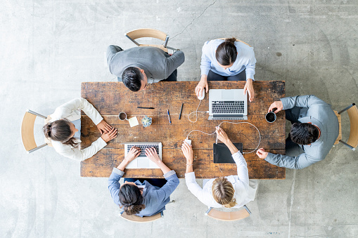 A group of small business professionals gather around a wooden desk as they meet together to discuss the future of the business.  They are each dressed professionally in suits and have laptops and reference materials out in front of them as they work collaboratively.