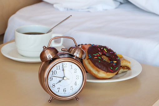 Stock photo showing close-up view of a cup of freshly brewed coffee, in white cup and saucer, on night stand with a plate containing a glazed, ring doughnut and a muffin in a paper cake case besides a retro, double bell, analogue alarm clock.