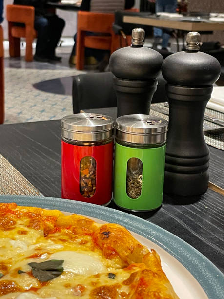 imagen de la escena del restaurante de la mesa del comedor de la pizzería con molinillos de sal y pimienta de madera negra, ollas cocteleras de hierbas secas y hojuelas de chile, pizza margherita en el plato, juego de cruet, enfoque en primer plano - pepper mill pepper shaker rock salt fotografías e imágenes de stock