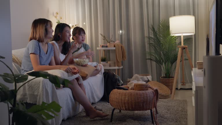 Young Asian women friends on sofa eat popcorn funny laugh watch comedy channel movie or sitcoms show on television in streaming online in home at night. Leisure activity concept.