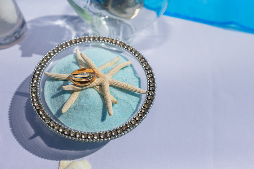 Diamond and gold rings in wedding ceremonies stacked on top of 2 starfish ornaments in glass, inside with blue sand surrounded by diamonds, very beautiful and outstanding
