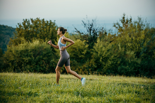 Side view of a sporty woman running on the grassy mountain during the summer sunset.