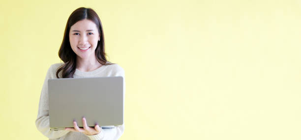 istock Young asian woman holding laptop computer, smiling and looking at camera while standing over isolated yellow background with copy space 1455654417