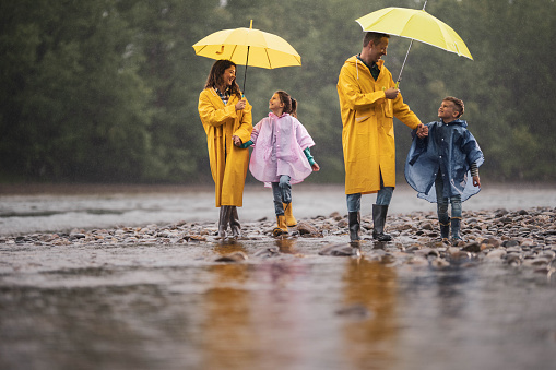 Happy family in raincoats enjoying in talk and walk on a rainy day at riverbank. Parents are holding umbrellas.