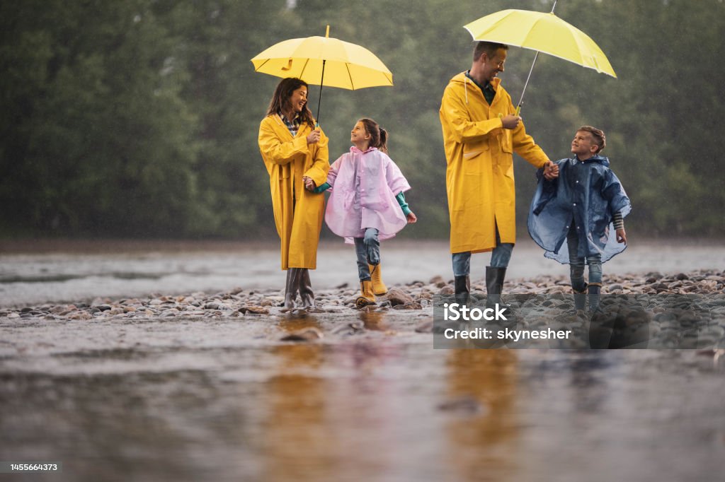 Carefree family in raincoats walking by the river on a rainy day. Happy family in raincoats enjoying in talk and walk on a rainy day at riverbank. Parents are holding umbrellas. Rain Stock Photo