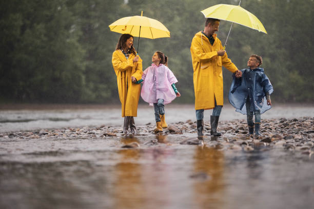 Carefree family in raincoats walking by the river on a rainy day. Happy family in raincoats enjoying in talk and walk on a rainy day at riverbank. Parents are holding umbrellas. weather protection clothing stock pictures, royalty-free photos & images