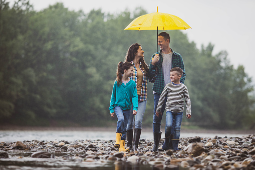 Happy parents and their small kids talking while walking on a stone beach by the river during rainy day. Copy space.