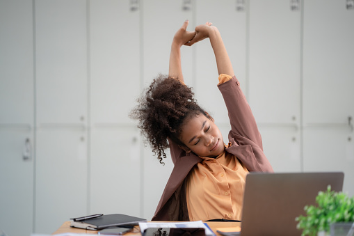 Young African American businesswoman stretching with hand raised while sitting on a chair in an office.