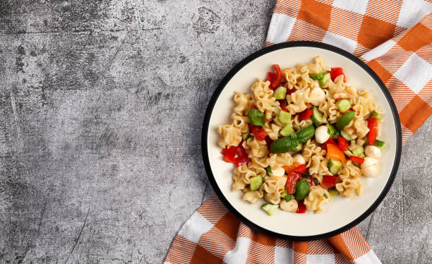 istock Pasta salad with mozzarella, greens, cucumbers and tomatoes on a round plate on a dark gray background. Top view, flat lay 1455667904