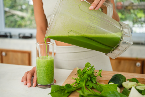 An unrecognizable woman is pouring a green smoothie into a glass.