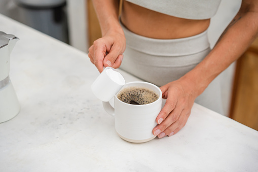 An unrecognizable woman is adding a protein powder in a cup of coffee.