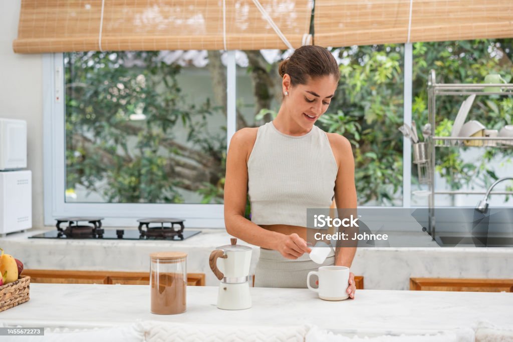 Smiling woman adding protein powder in a coffee Smiling woman is adding a protein powder in a cup of coffee in the kitchen after workout. Protein Drink Stock Photo