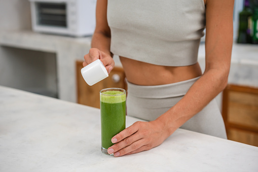 An unrecognizable woman is adding a protein powder in a glass of green smoothie.