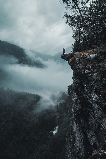 A vertical drone shot of a man on the edge of a rocky cliff facing the foggy forests