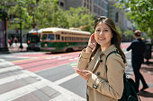 cheerful asian Taiwanese girl tourist tucking hair in breeze and smiling at camera with phone in hand at roadside with two city buses stopping at background in san Francisco California usa