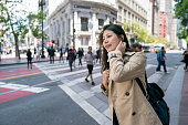 asian Korean female businessperson tucking hair with smile and looking into distance while waiting for bus near crosswalk in san Francisco California usa. people are crossing road at background