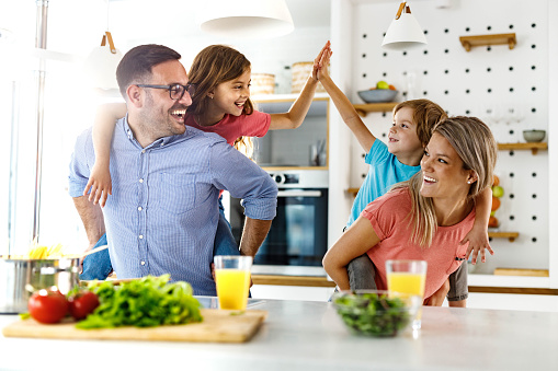 Happy parents having fun while piggybacking their kids in the kitchen who are giving each other high-five.