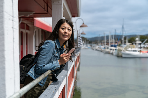 excited asian chinese girl holding phone and craning neck to look into space while enjoying scenic view from waterfront railing near fishing fleet port at Old Fisherman's Wharf in usa