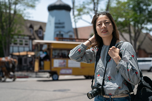 portrait of asian japanese woman tourist carrying camera and admiring beautiful townscape of solvang with giant windmill and people having horse drawn trolley tour at background in California