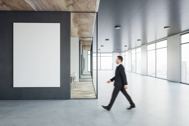 istock Walking businessman by blank white poster on black wall background in sunlit modern interior design office with concrete floor, meeting room with wooden decoration and city view background, mock up 1455682475