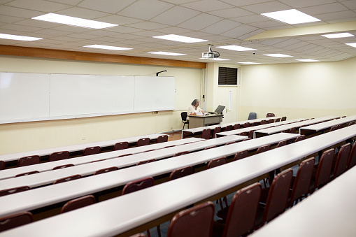 View across tiered rows of unoccupied student desks and chairs as mature woman sits at desk, front of room, and looks at paperwork. Property release attached.
