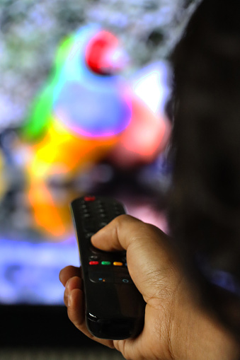 Stock photo showing a black TV remote control being operated by the hand of an unrecognisable person pressing the buttons and changing the channels, pointing the control at the television screen.