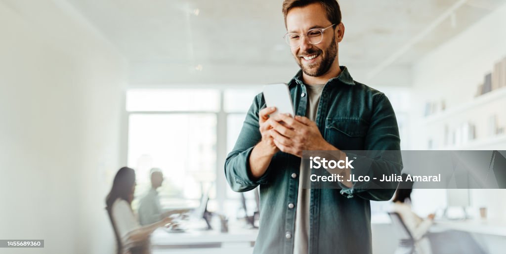 Business man reading a message on a mobile phone in an office Business man reading a message on a mobile phone in an office. Male business professional standing in a co-working office. Mobile Phone Stock Photo