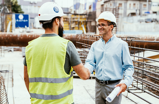 Two men engineers in workwear shaking hands against construction site, close up