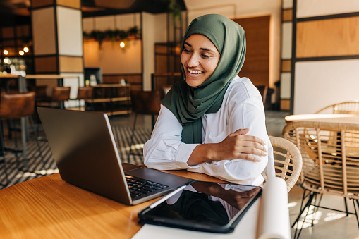 Muslim university student smiling happily while having a video conference with her tutor in a cafe. Cheerful woman with a hijab attending an online class in a coffee shop.
