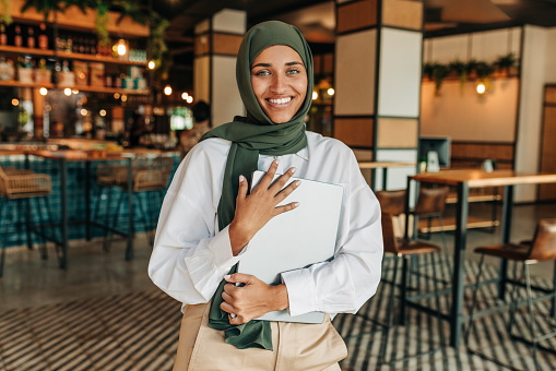 Happy young Muslim woman smiling at the camera while standing in a cafe. Portrait of a young freelancer with a hijab holding a laptop in a coffee shop.
