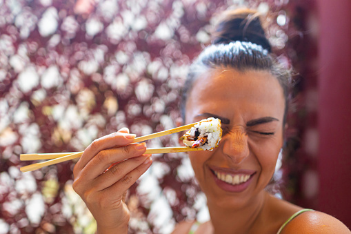 Close-up shot of a beautiful happy Caucasian woman holding an Uramaki roll with chopsticks and making a face at the sushi bar.