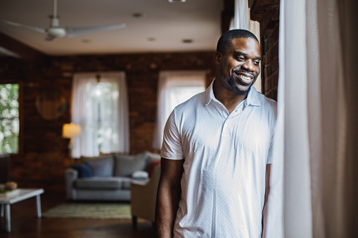 Copy space shot of charming Black man, in white t-shirt, leaning against the brick wall, by the window, looking outside, smiling and contemplating.