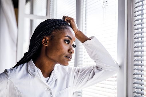 Portrait of overworked female scientist standing by the window, head in hand, looking outside, pondering while taking a break from doing research at the laboratory.