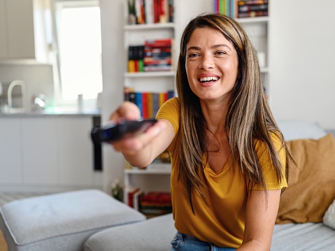 Shot of a young woman  holding the remote control and watching tv at home