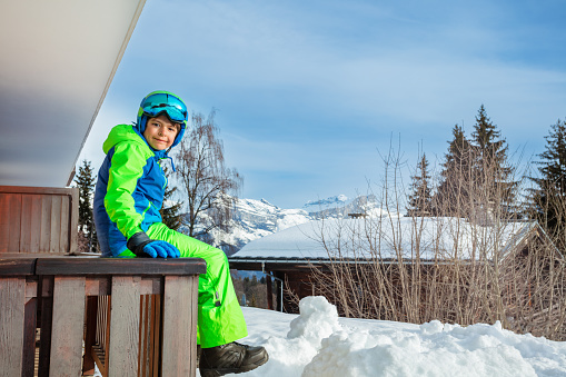 Cute child sit on balcony rail smiling in ski outfit glasses and helmet with mountain peak on background