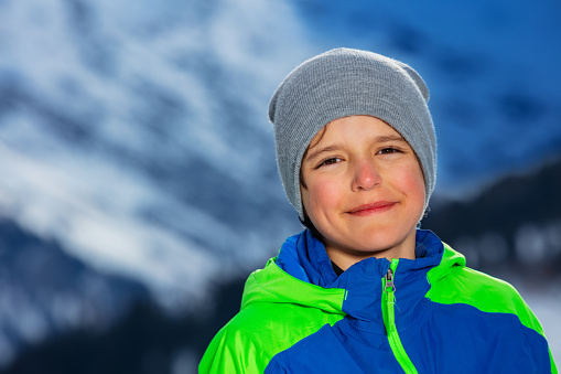 Smiling boy stand wearing wither coat and hat over snowy winter mountain slopes
