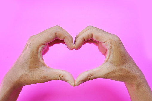 asian man hand forming a heart shape using the index finger and thumb, love symbol on pink background.