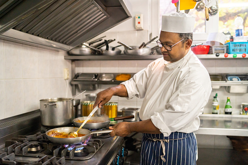 An Indian restaurant kitchen worker is creating and stirring a curry sauce in a cooking pan on a commercial cooker