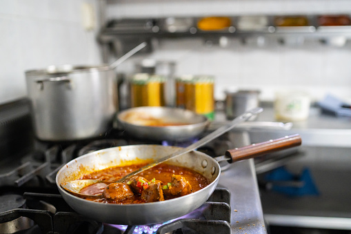 Close up of a curry in a frying pan on a lit gas flame of a commercial stove