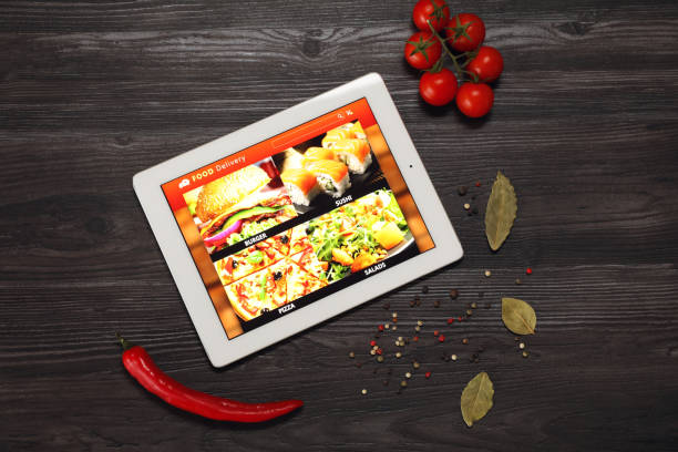 istock Modern tablet with open page for online food ordering surrounded by scattered products on wooden table, flat lay. Delivery service 1455707017