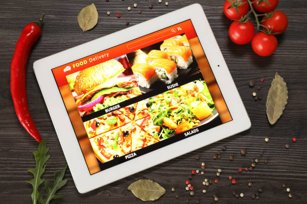 istock Modern tablet with open page for online food ordering surrounded by scattered products on wooden table, flat lay. Delivery service 1455707693