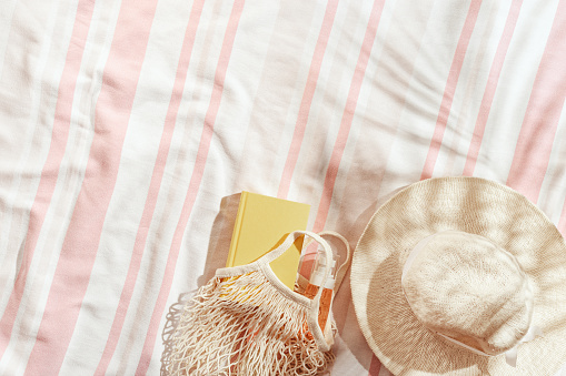 Summer background, top view wide-brimmed sun hat, cosmetics products, book on striped beach towel, pale pink background, sun shadows from palm leaf. Summer pastel color aesthetic photo, copy space