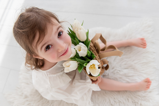 Top view close-up portrait of little girl in a white dress, nightgown. Toddler embraces a bouquet of fresh, delicate white tulips. Gift for the holiday, the concept of purity, spring time. Copy space