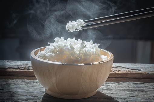The man hand of using black chopsticks holding hot jasmine rice with smoke and steam in black bowl over dark background.hot food concept
