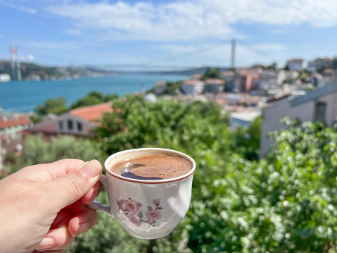 closeup female hand holding cup of Turkish coffee over sea view in Istanbul, Turkey or Turkiye. hand holding cup of hot coffee over Bosphorus bridge under blue sky in Istanbul, Turkey with copy space