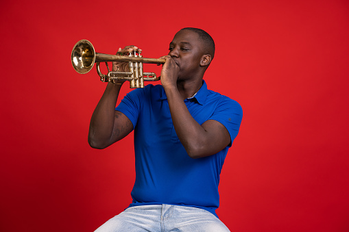 African american man sitting on a chair and plays the trumpet in front of a red background