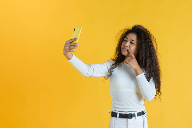 Young woman taking selfie smiling and touching her mouth against yellow background stock photo