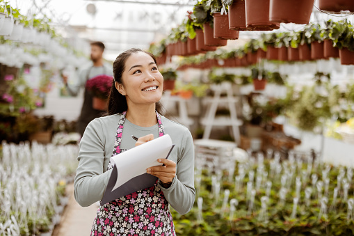Young Gardener Woman Taking Notes Holding a Clipboard, While Working in a Greenhouse. Woman Working in a Garden Center and Taking Notes of Her Plants