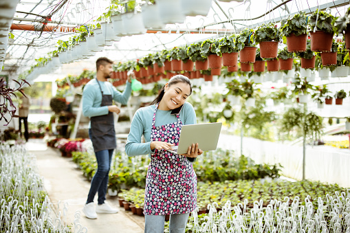 Asian businesswoman talking on cellphone while standing in apron in small floral center. Joyful female florist calling on smartphone at work. Own business concept
